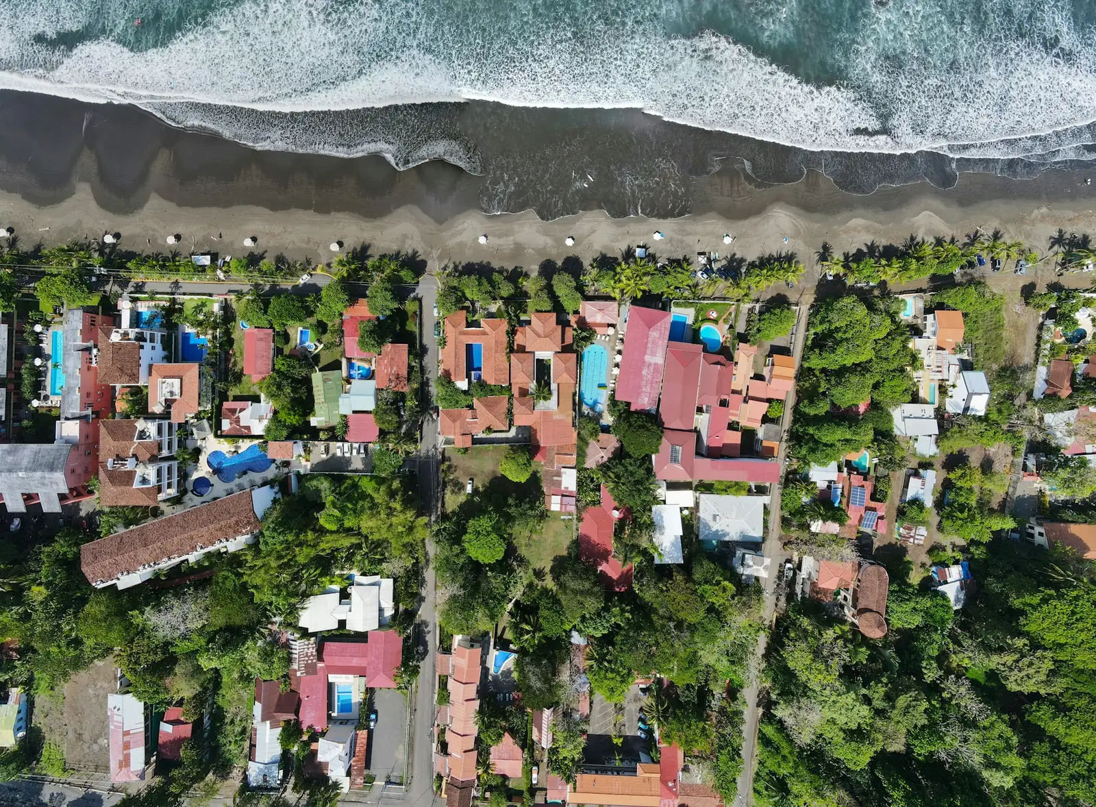 Aerial view of Jaco, Costa Rica, with hotels and palms lining the gray-sand beach as surf rolls in