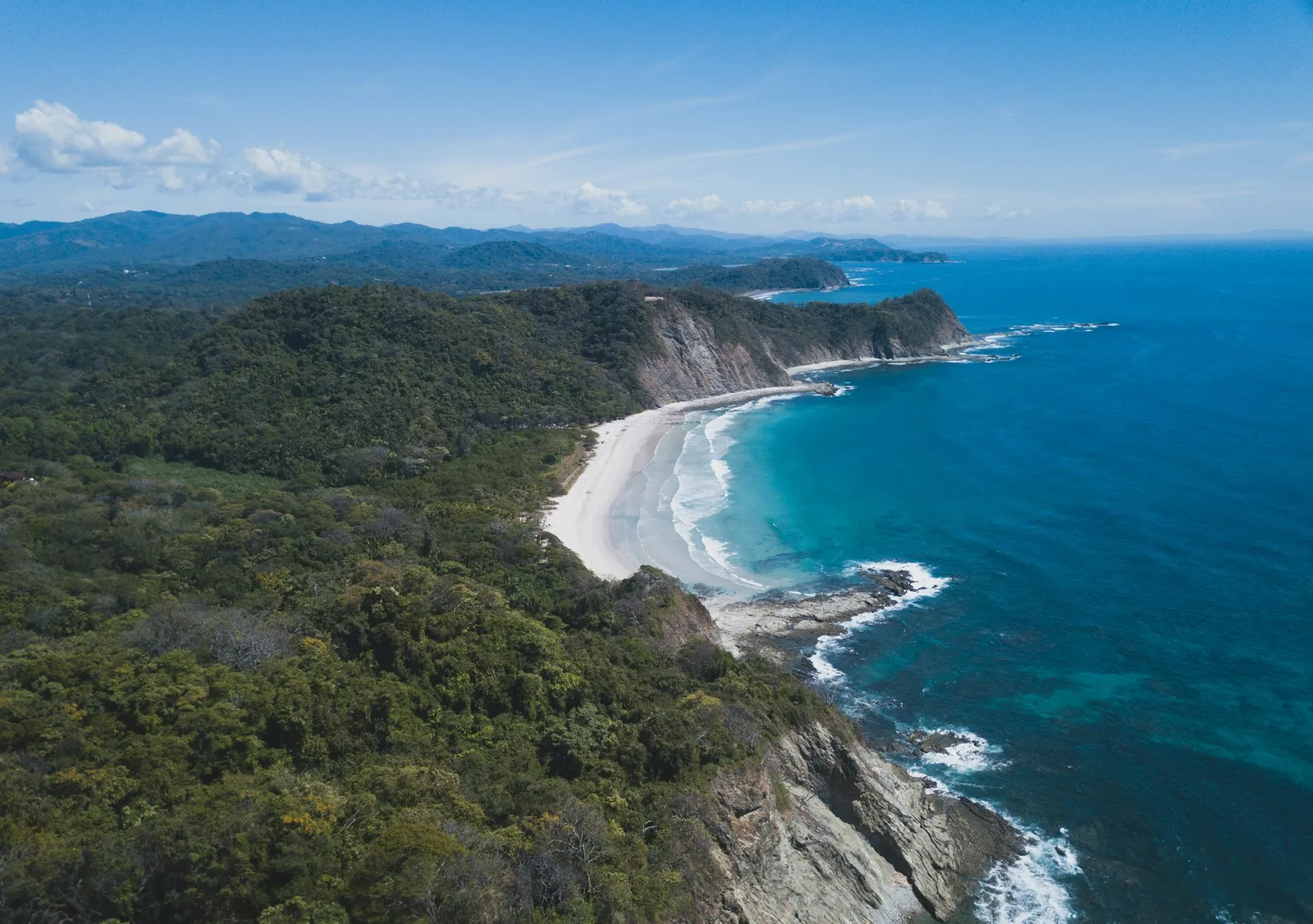 Aerial view of a forested headland wrapping a curved white-sand cove and turquoise Pacific water on the Guanacaste coast of Costa Rica